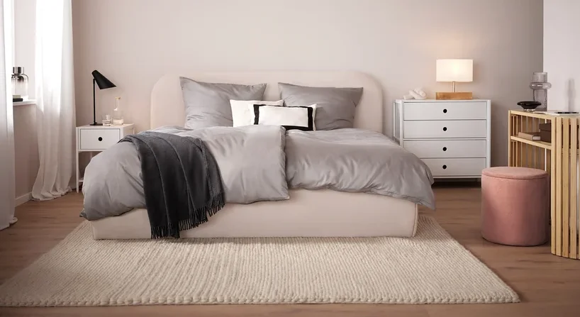 Schlafzimmer mit beigem Bett, grauer Bettwäsche und beigem Teppich. Im Hintergrund weißer Nachttisch und Kommode.