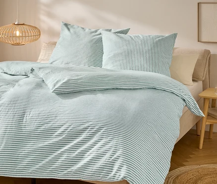 Bett mit grün-weiß gestreifter Bettwäsche und zwei Kissen. Nachttisch mit Deko und Lampe hängen von der Decke.