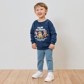 Ein lächelnder Junge trägt ein blaues Paw Patrol-Sweatshirt, Jeans und weiße Turnschuhe.
