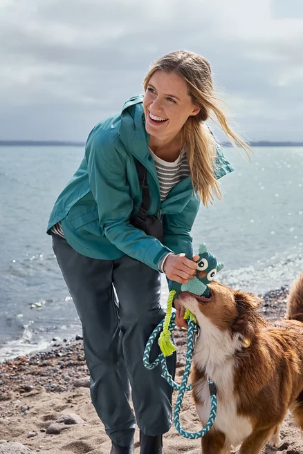 Eine lächelnde Frau in türkiser Jacke spielt am Strand mit einem braun-weißen Hund Zerrspiel mit einem grün-blauen Plüschtier. Im Hintergrund ist Wasser unter bewölktem Himmel.