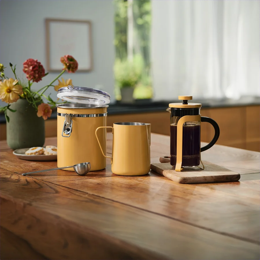 Ein gelber Behälter mit offenem Deckel, ein gelber Metallbecher und ein silberner Messlöffel stehen auf einem Holztisch. Rechts davon steht eine French Press mit Kaffee auf einem kleinen Schneidebrett. Eine Blumenvase und Kekse auf einem Teller befinden sich links.