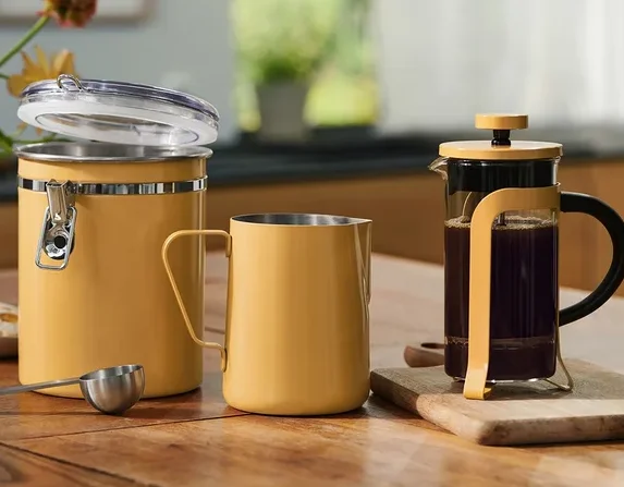 Ein gelber Behälter mit offenem Deckel, ein gelber Metallbecher und ein silberner Messlöffel stehen auf einem Holztisch. Rechts davon steht eine French Press mit Kaffee auf einem kleinen Schneidebrett. Eine Blumenvase und Kekse auf einem Teller befinden sich links.