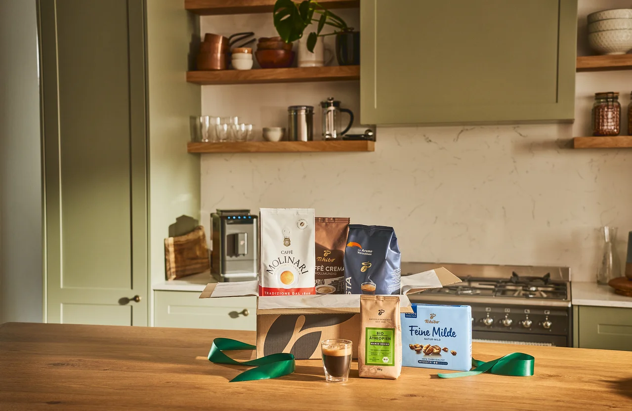 Verschiedene Kaffeepackungen in einer offenen Schachtel auf einem Holztisch, zusammen mit einem Glas Espresso und einem grünen Band.