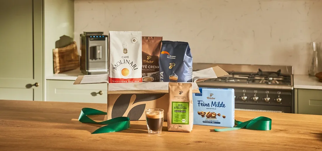 Verschiedene Kaffeepackungen in einer offenen Schachtel auf einem Holztisch, zusammen mit einem Glas Espresso und einem grünen Band.