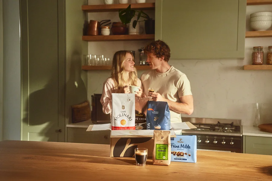 Zwei Personen in einer Küche, ein Mädchen hält eine Tasse, ein Mann hält ein kleines Glas, und vor ihnen steht eine offene Kiste mit Molinari- und Tchibo-Kaffees.