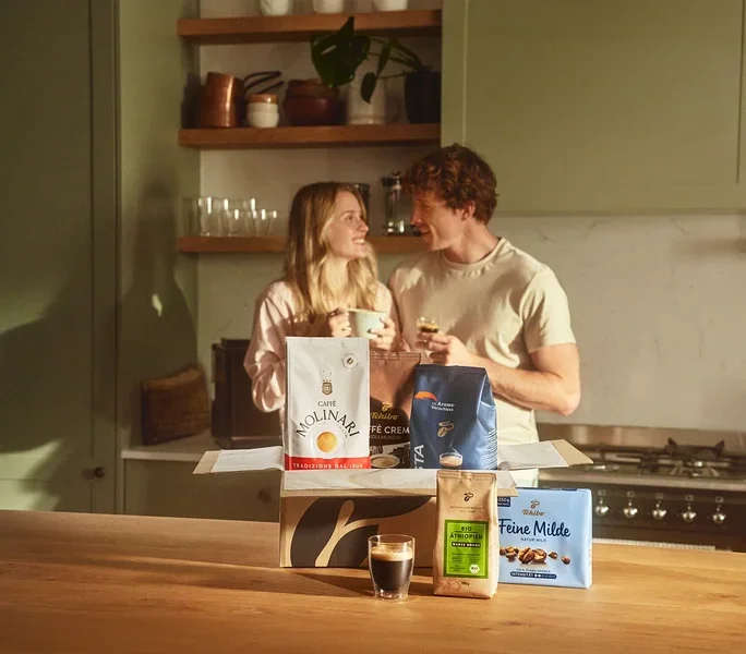Zwei Personen in einer Küche, ein Mädchen hält eine Tasse, ein Mann hält ein kleines Glas, und vor ihnen steht eine offene Kiste mit Molinari- und Tchibo-Kaffees.