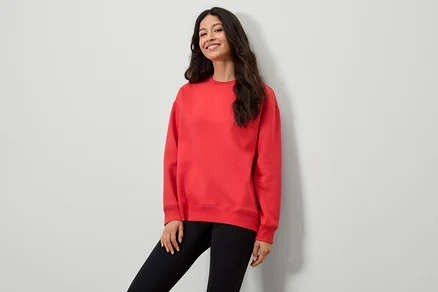 Frau in rotem Sweatshirt und schwarzen Leggings posiert vor einer weißen Wand.