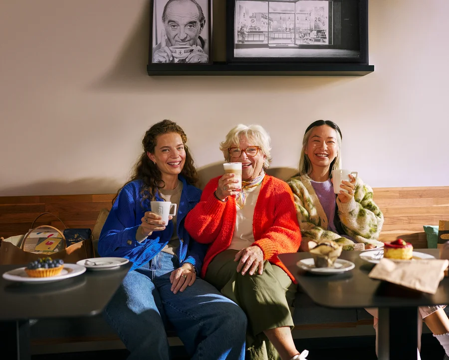 Drei Frauen unterschiedlichen Alters sitzen lächelnd auf einer Bank in einem Café, trinken Kaffee und essen Gebäck. Eine Frau hält eine Tasse, eine andere ein Latte-Glas und die dritte eine Tasse.