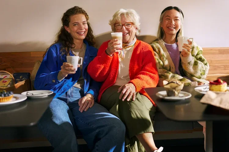 Drei Frauen unterschiedlichen Alters sitzen lächelnd auf einer Bank in einem Café, trinken Kaffee und essen Gebäck. Eine Frau hält eine Tasse, eine andere ein Latte-Glas und die dritte eine Tasse.