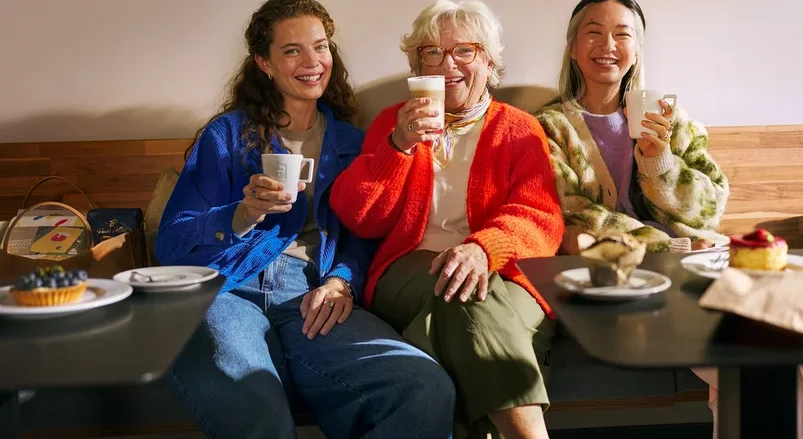 Drei Frauen unterschiedlichen Alters sitzen lächelnd auf einer Bank in einem Café, trinken Kaffee und essen Gebäck. Eine Frau hält eine Tasse, eine andere ein Latte-Glas und die dritte eine Tasse.