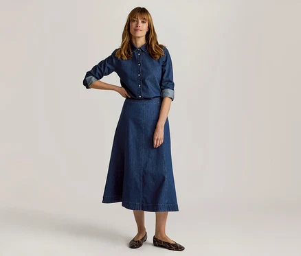 Frau in Jeanshemd und A-Linien-Midirock, mit einer Hand in die Hüfte gestemmt, trägt gemusterte flache Schuhe.