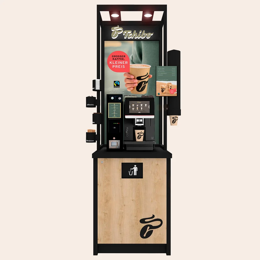 Eine schwarze und holzfarbene Tchibo Selbstbedienungs-Kaffeemaschine mit einem Bildschirm, der Kaffeeoptionen und eine Hand mit einem Kaffeebecher zeigt. Unten ist ein Mülleimer-Symbol.