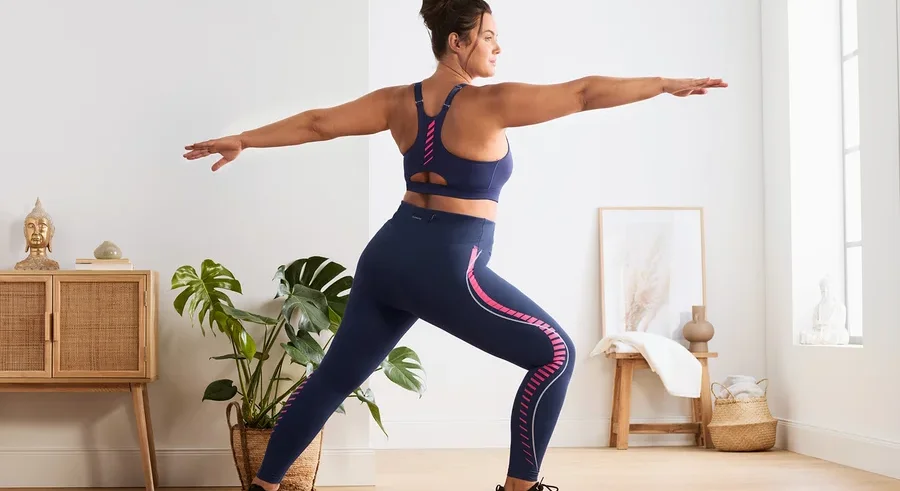 Eine Frau in einem marineblauen Sport-BH mit Frontreißverschluss und Sporttight steht in Krieger-II-Pose.