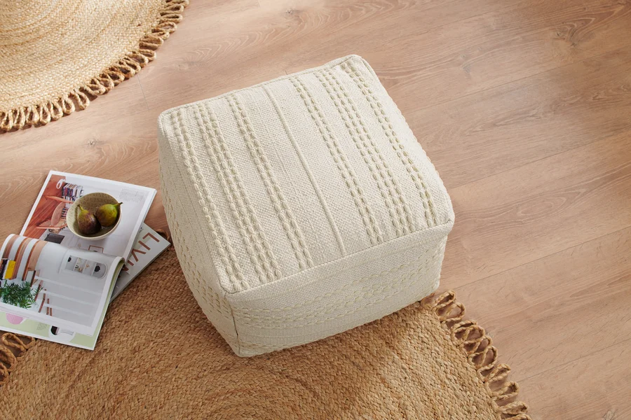 Ein cremefarbener, gestrickter Sitzpouf auf einem Holzboden, daneben liegen Zeitschriften und eine Schale mit Obst.