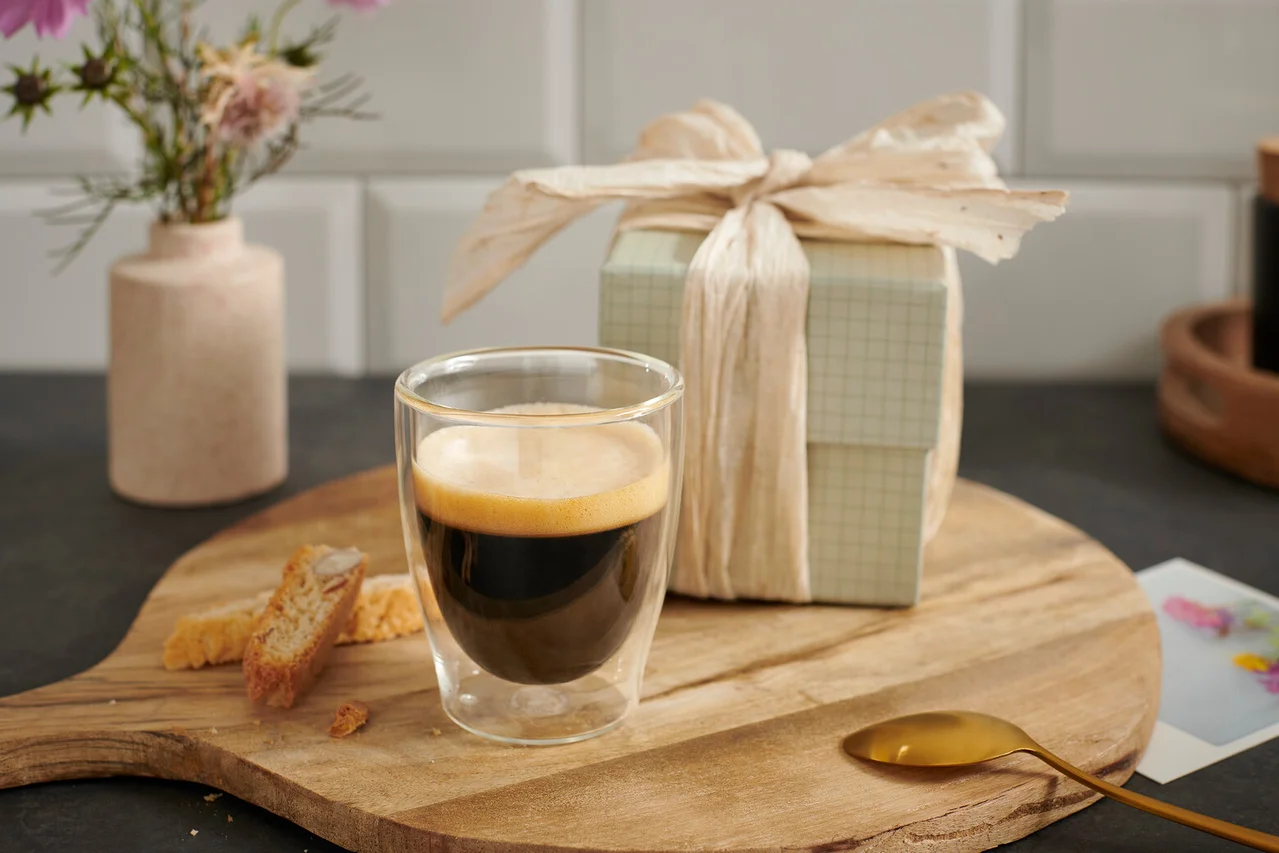Eine Tasse Espresso auf einem Holzbrett neben einem Geschenk und einer Blume.