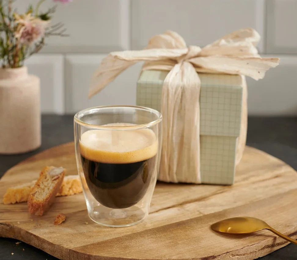 Eine Tasse Espresso auf einem Holzbrett neben einem Geschenk und einer Blume.