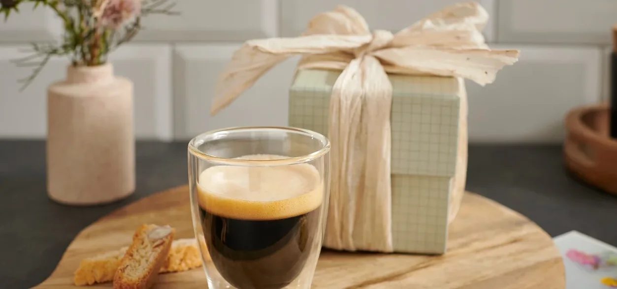 Eine Tasse Espresso auf einem Holzbrett neben einem Geschenk und einer Blume.