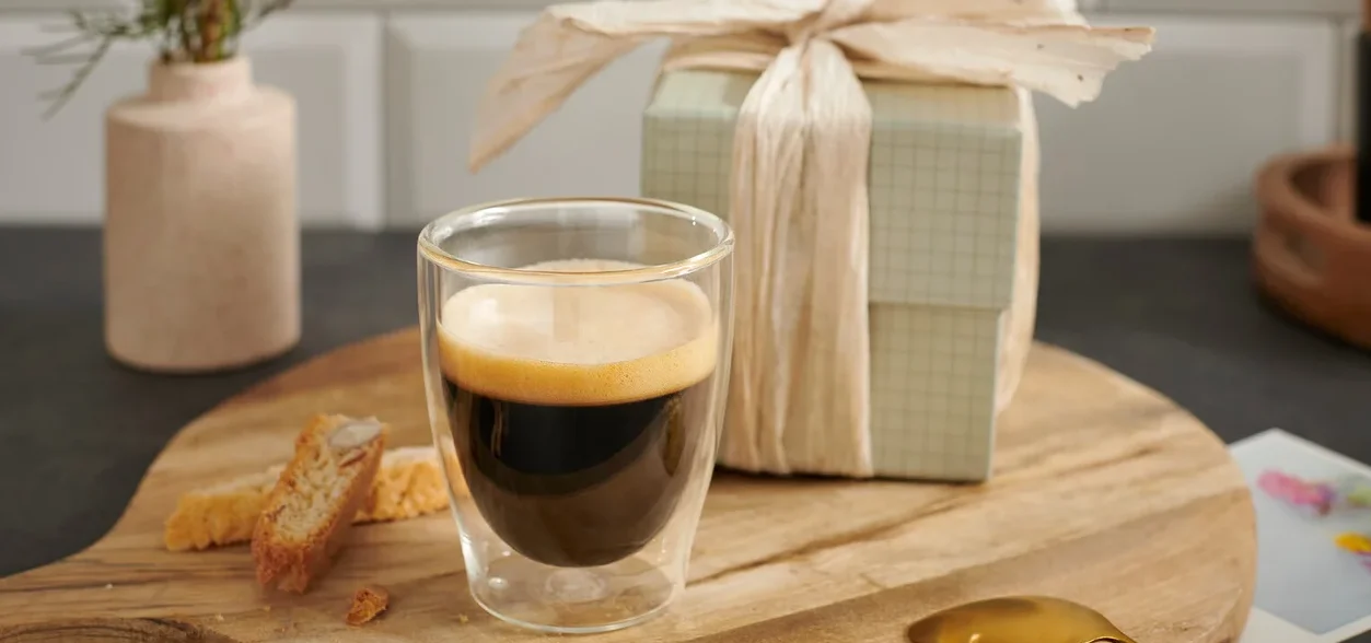 Eine Tasse Espresso auf einem Holzbrett neben einem Geschenk und einer Blume.