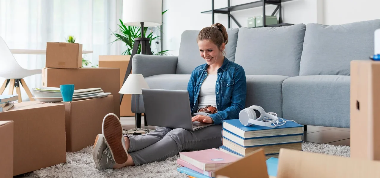Frau sitzt auf dem Boden, umgeben von Kisten, und benutzt einen Laptop. Im Hintergrund befindet sich ein graues Sofa.