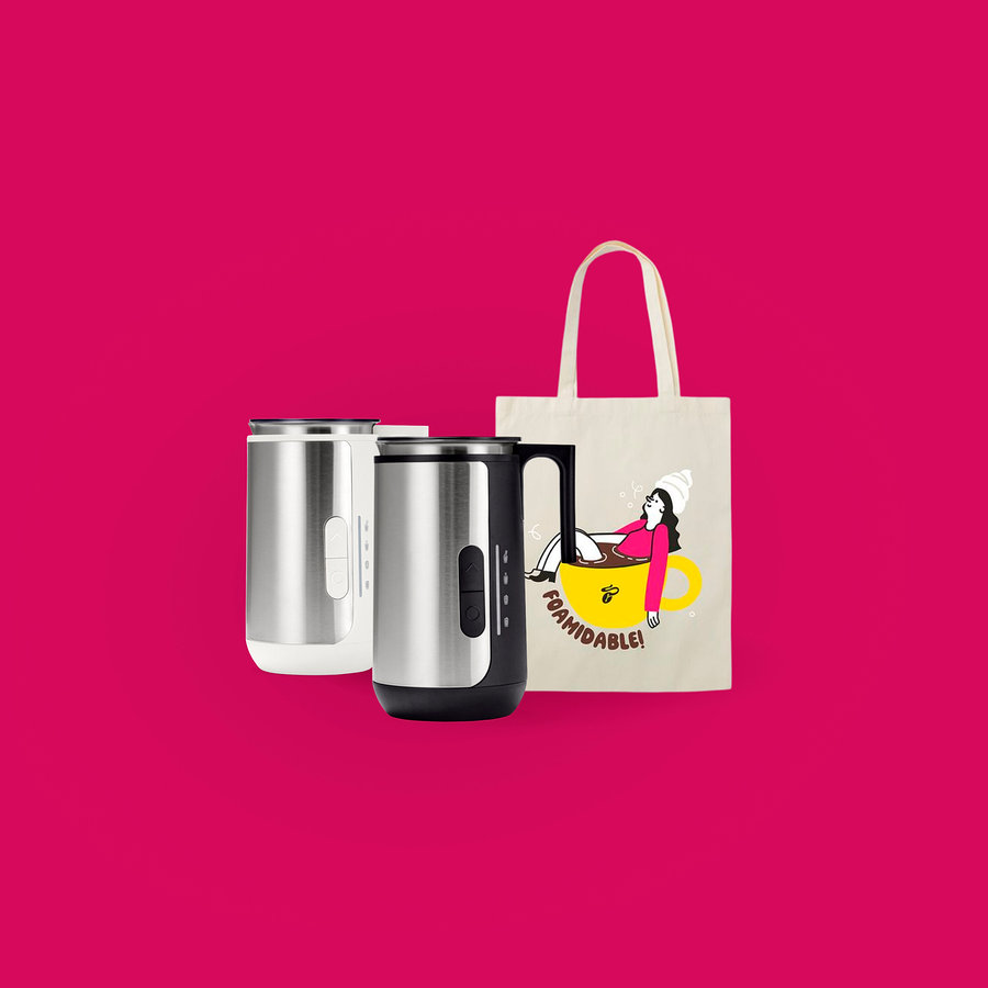 Zwei Milchaufschäumer in Silber und Weiß stehen neben einer Tragetasche mit dem Aufdruck einer Frau, die in einer gelben Tasse sitzt, auf einem pinkfarbenen Hintergrund.