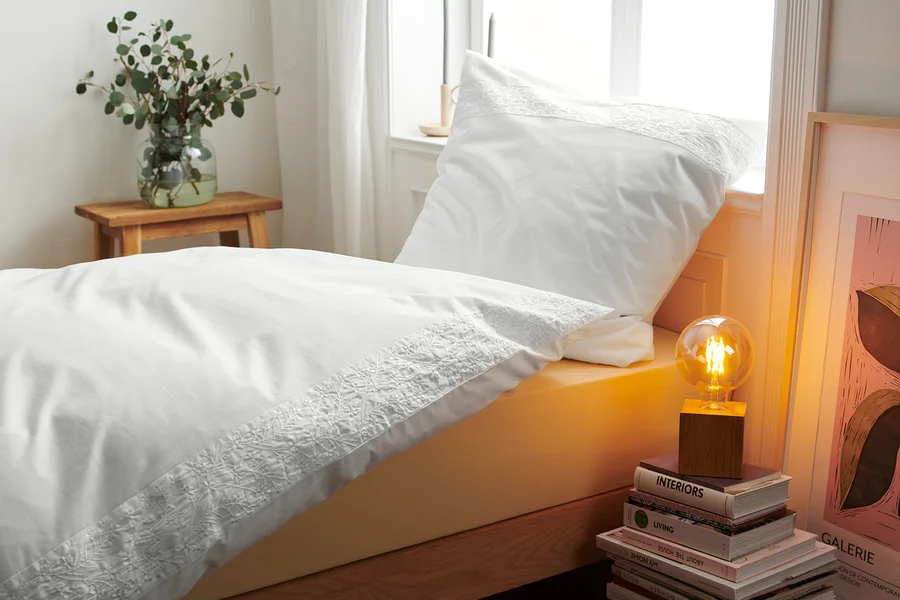 Detailansicht von weisser bestickter Renforcé-Bettwäsche auf einem Bett neben einer Lampe und Büchern.