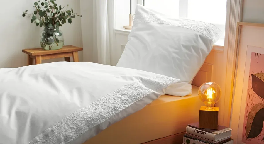 Detailansicht von weisser bestickter Renforcé-Bettwäsche auf einem Bett neben einer Lampe und Büchern.