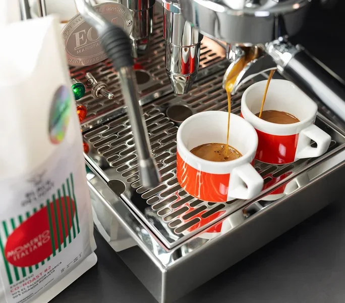 Kaffee läuft in zwei rote Tassen aus einer Kaffeemaschine neben einer Packung Black Delight gemahlenem Kaffee.