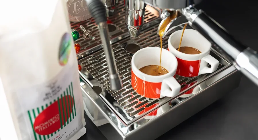 Kaffee läuft in zwei rote Tassen aus einer Kaffeemaschine neben einer Packung Black Delight gemahlenem Kaffee.