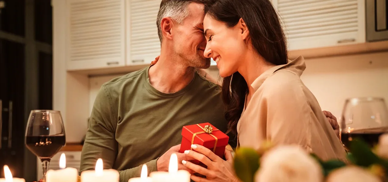 Ein lächelndes Paar umarmt sich an einem Tisch mit Kerzen und hält eine rote Geschenkbox.