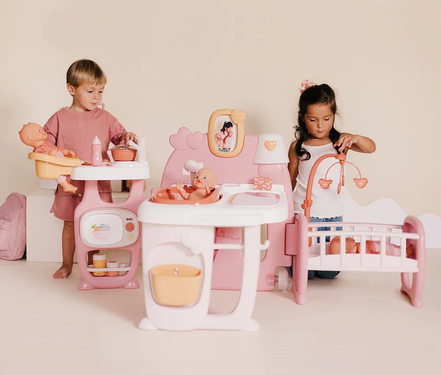 Zwei Kinder spielen mit einem Smoby Baby Nurse Puppen-Spielcenter, das eine Puppenküche, ein Bett und eine Badestation umfasst.