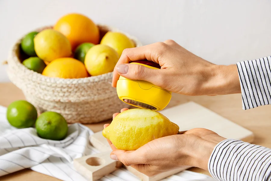 Eine Person schält eine gelbe Zitrone mit einem gelben Schäler. Im Hintergrund befindet sich ein Korb mit Zitronen, Limetten und Orangen.