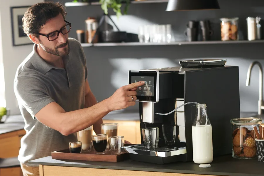 Mann bedient Tchibo Office Kaffeevollautomat, schwarz. Auf dem Tisch stehen Kaffeetassen und Milch.