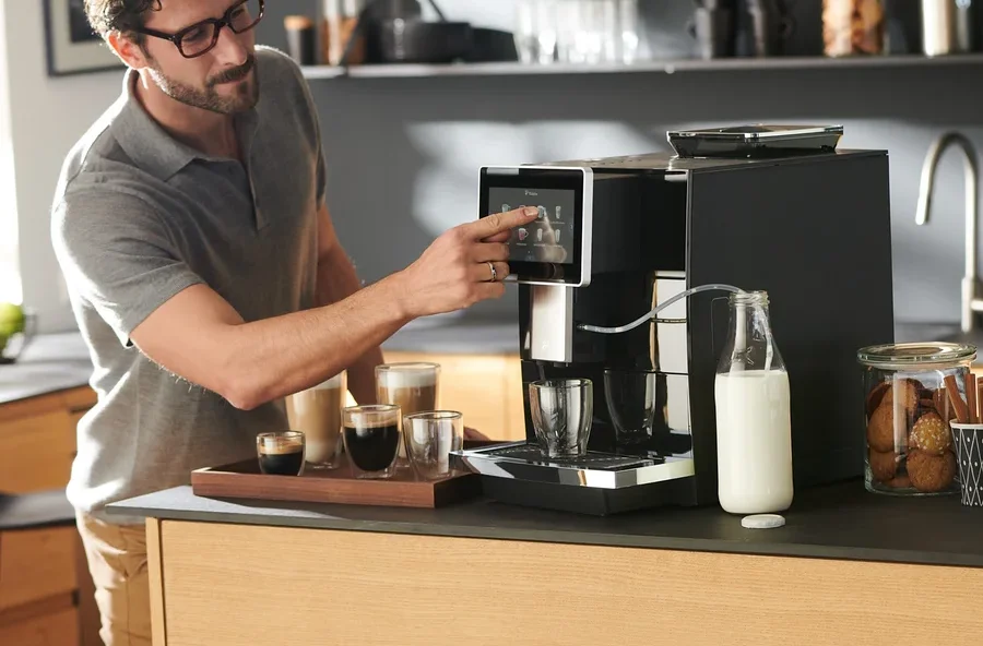 Mann bedient Tchibo Office Kaffeevollautomat, schwarz. Auf dem Tisch stehen Kaffeetassen und Milch.