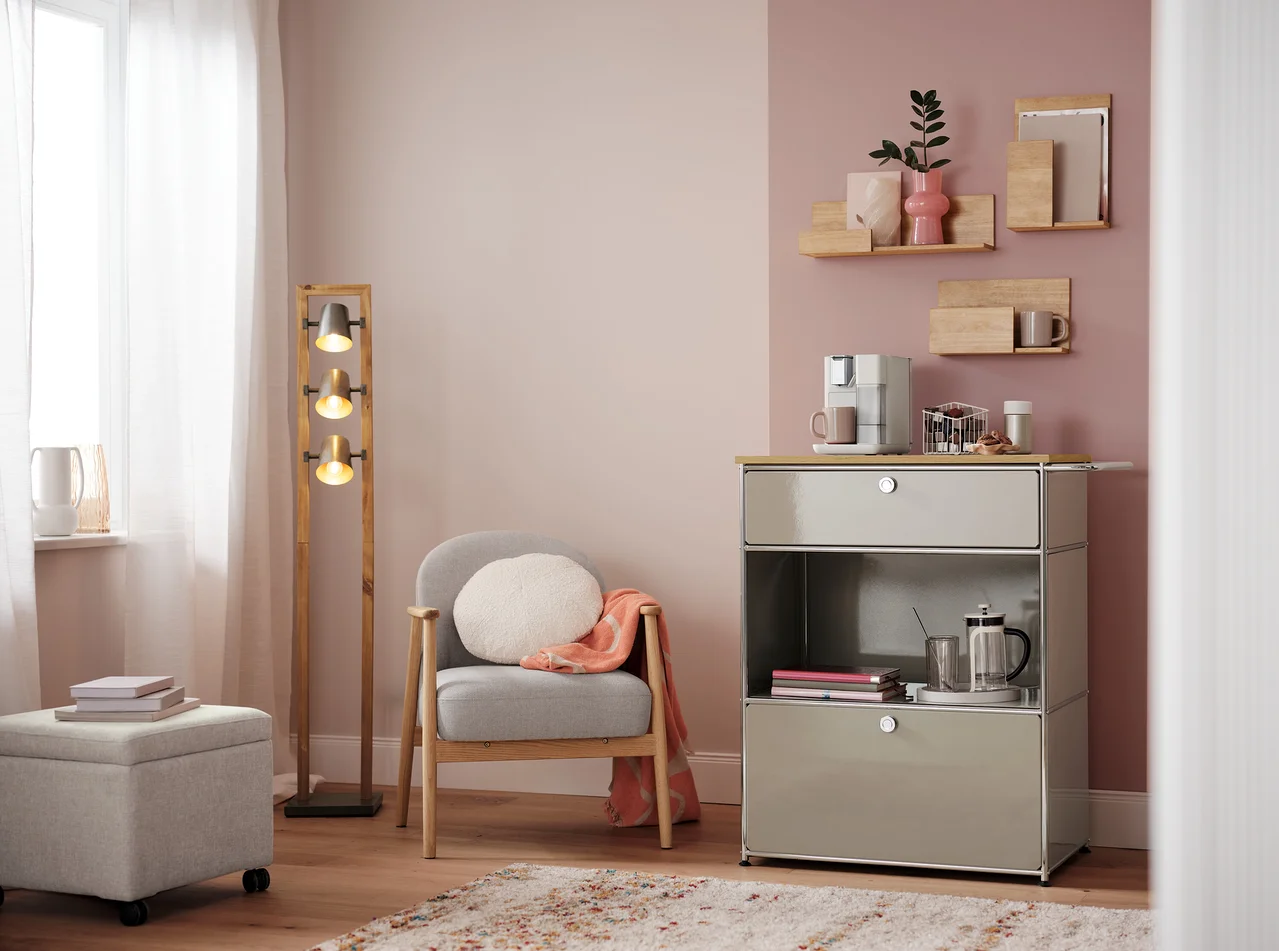 Wohnzimmer mit Sessel, Lampe und Kommode mit Kaffeemaschine. Auf der Kommode steht ein Kapselkorb, weiß.