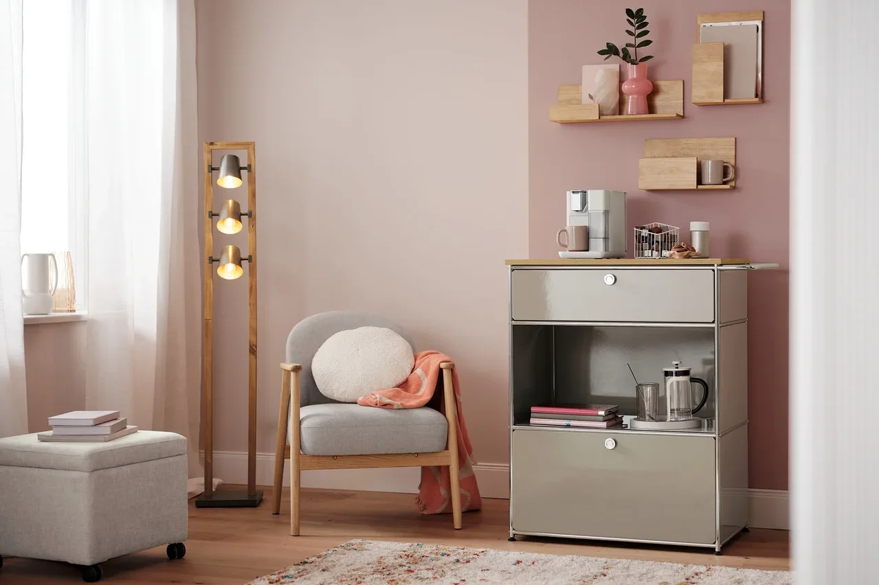 Wohnzimmer mit Sessel, Lampe und Kommode mit Kaffeemaschine. Auf der Kommode steht ein Kapselkorb, weiß.