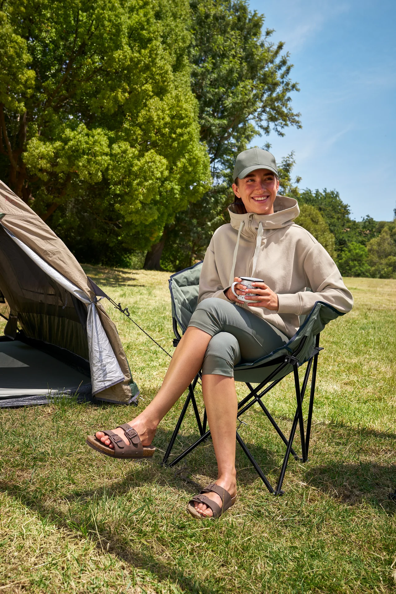 Frau sitzt im Tambu »ASANA« Camping-Faltstuhl, hält eine Tasse und trägt 3/4-Outdoor-Leggings und einen Kapuzenpullover.