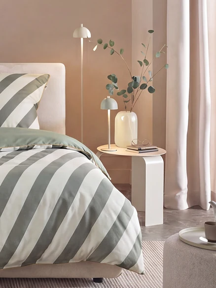 Bett mit grün-weiß gestreifter SCHÖNER WOHNEN-Kollektion Renforcé-Bettwäsche, daneben ein Beistelltisch mit Lampe, Vase und Brille, sowie eine Stehlampe.
