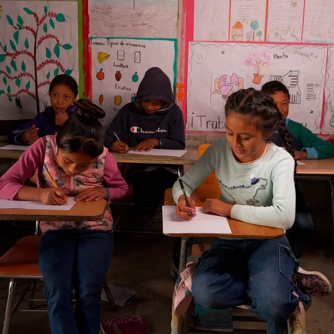 Mehrere Kinder sitzen in einem Klassenzimmer an Holztischen und schreiben auf Papier. Zwei Mädchen im Vordergrund konzentrieren sich auf das Schreiben, eines in einer rosa Weste und das andere in einem hellblauen Pullover. Die Wände des Klassenzimmers sind mit Lernplakaten dekoriert.