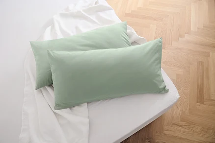 Zwei salbeigrüne Jersey-Kopfkissenbezüge liegen auf einem weißen Bettlaken.