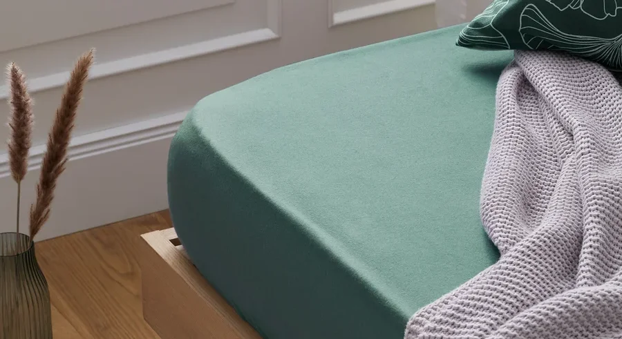 Grünes Biber-Spannbettlaken auf einem Bett mit einer Strickdecke darüber.