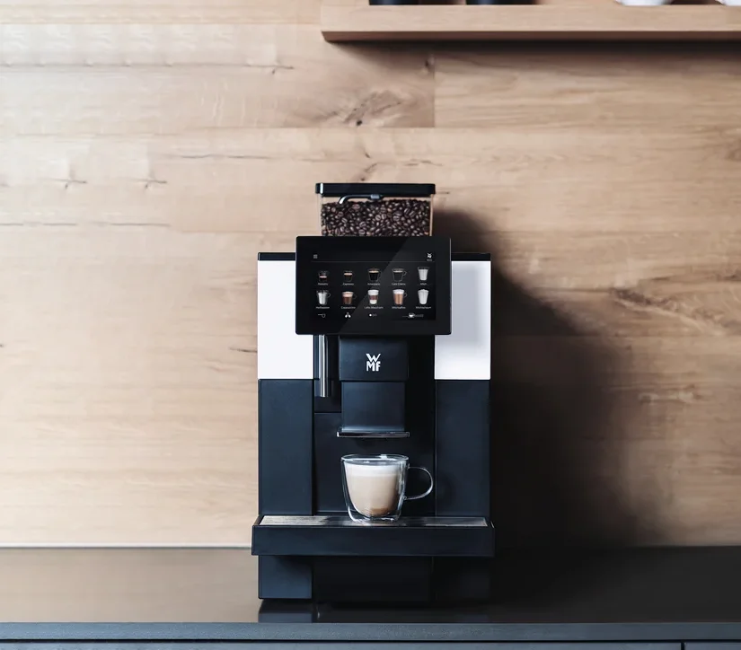 Profi-Kaffeevollautomat »WMF 950 S« vor einer Holzwand. Auf einem Regal darüber befinden sich zwei Blumentöpfe und Tassen.