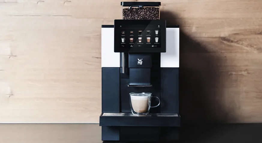 Profi-Kaffeevollautomat »WMF 950 S« vor einer Holzwand. Auf einem Regal darüber befinden sich zwei Blumentöpfe und Tassen.