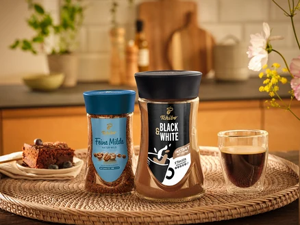 Zwei Gläser Tchibo Feine Milde und Black & White Instantkaffee auf einem geflochtenen Tablett mit einem Stück Schokoladenkuchen und einer Tasse Kaffee.