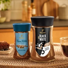 Zwei Gläser Tchibo Feine Milde und Black & White Instantkaffee auf einem geflochtenen Tablett mit einem Stück Schokoladenkuchen und einer Tasse Kaffee.