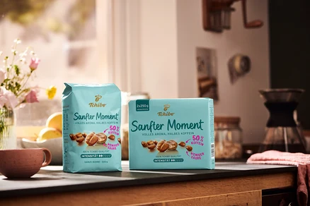 Zwei Packungen Tchibo Sanfter Moment Kaffee mit 50 % Koffein stehen auf einem Tisch neben einer Tasse und einer Vase mit Blumen.