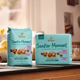 Zwei Packungen Tchibo Sanfter Moment Kaffee mit 50 % Koffein stehen auf einem Tisch neben einer Tasse und einer Vase mit Blumen.