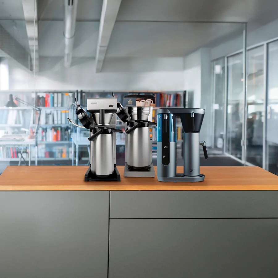 Drei Kaffeemaschinen stehen auf einer Holzplatte, im Hintergrund eine Bibliothek und Glaswände eines Büros.