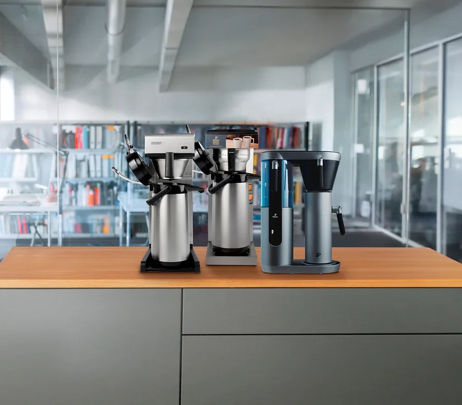 Drei Kaffeemaschinen stehen auf einer Holzplatte, im Hintergrund eine Bibliothek und Glaswände eines Büros.