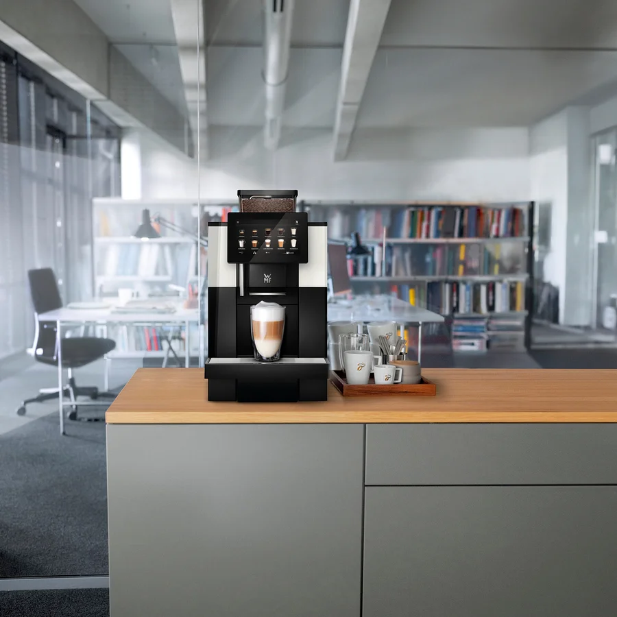 Ein Kaffeevollautomat steht auf einer Küchentheke mit einer Tasse Latte Macchiato davor. Daneben stehen Tassen und Gläser auf einem Holztablett.