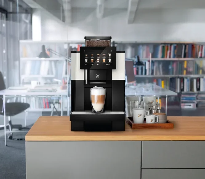 Ein Kaffeevollautomat steht auf einer Küchentheke mit einer Tasse Latte Macchiato davor. Daneben stehen Tassen und Gläser auf einem Holztablett.